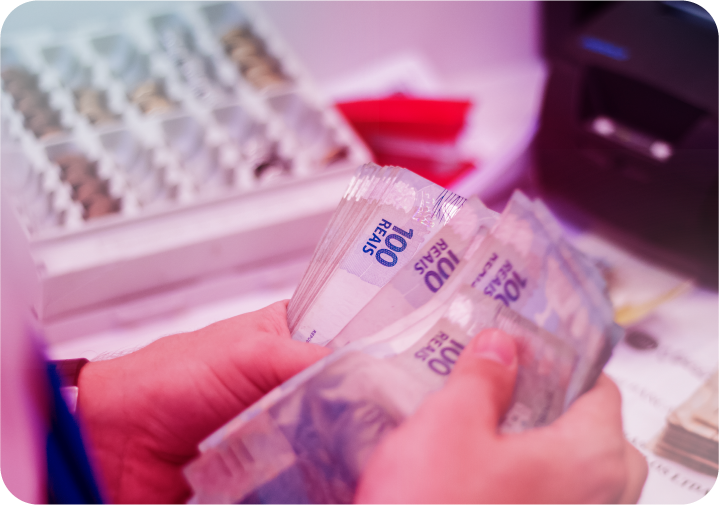 Pessoa contando cédulas de 100 reais com gaveta registradora ao fundo.   