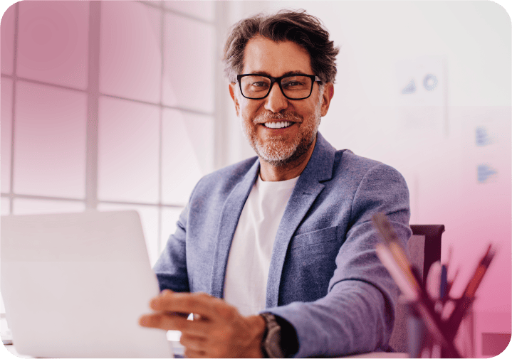 Homem sorridente usando óculos e blazer azul, trabalhando em notebook em ambiente iluminado.  