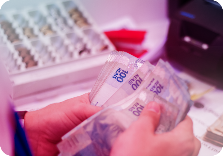 Pessoa contando cédulas de 100 reais com gaveta registradora ao fundo.   